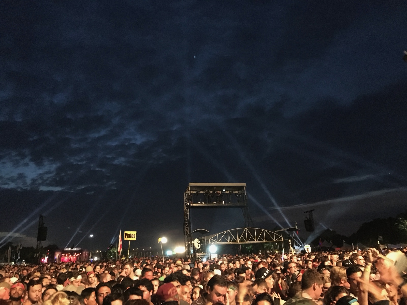 A huge crowd at night in front of a festival stage with spotlight beams in the sky